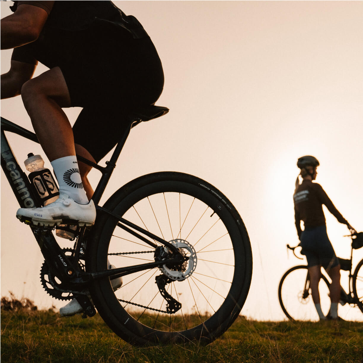Two cyclists are seen in a scenic outdoor setting during sunset, one riding and the other standing beside their bike.
